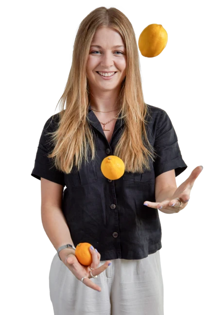 Jess is wearing black trousers and a beige cardigan as she sits on a wooden stool and looks directly into the camera with a big smile, her head slightly tilted.